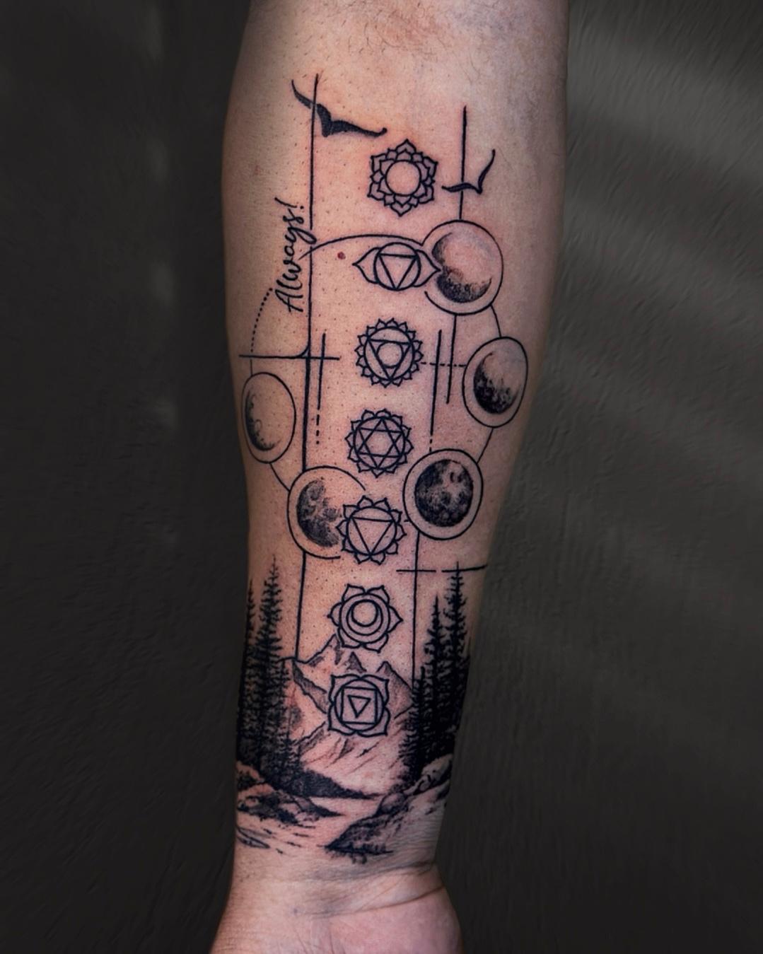 A forearm tattoo featuring a combination of celestial and natural elements, including phases of the moon, chakra symbols, and a scenic mountain landscape with trees.